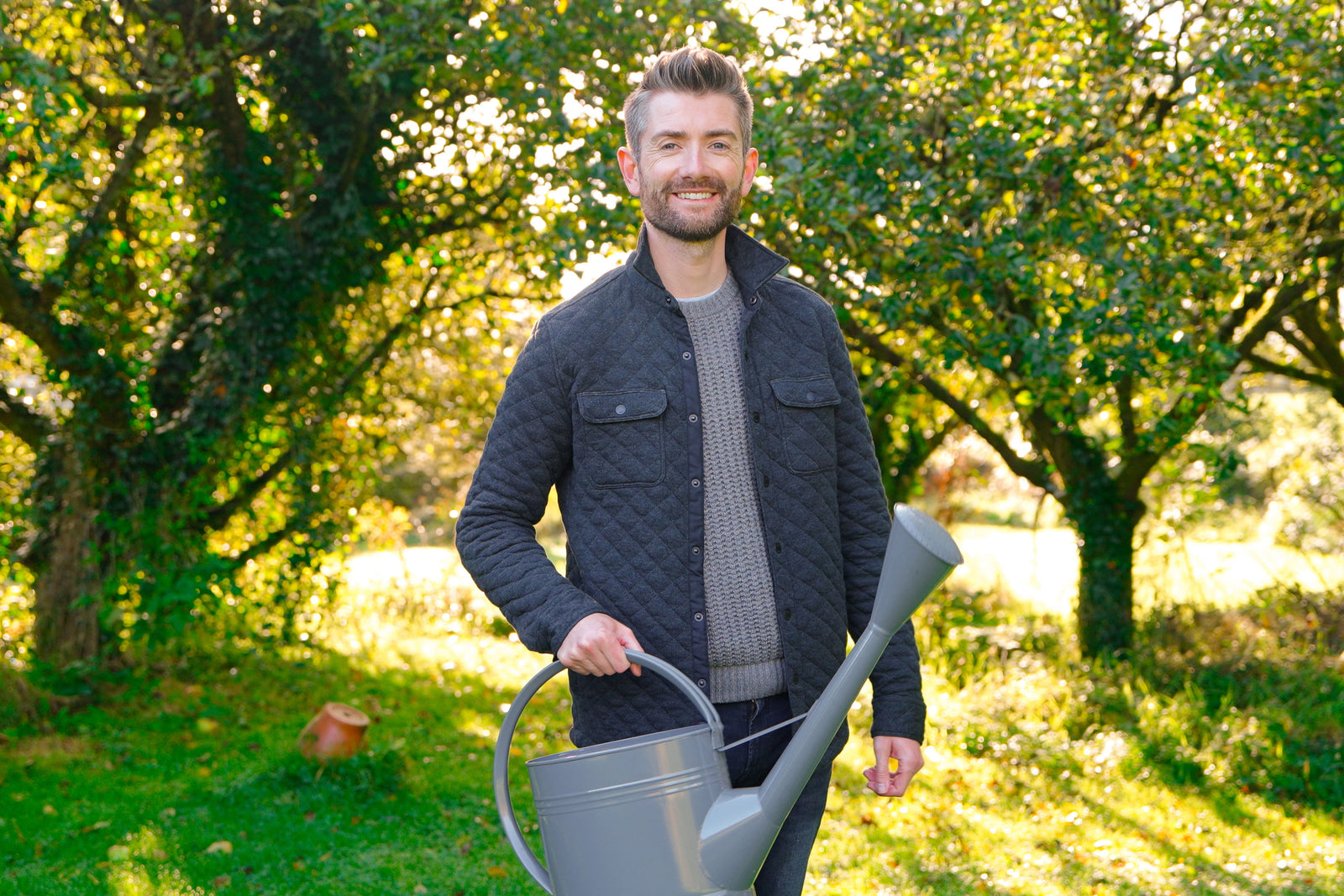 Niall stood in front of trees, wearing a grey padded jacket and holding a metal watering can