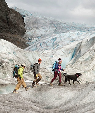 3 people wearing cold weather clothing, backpacks and Xtratuf boots with a black dog walking along a mountain ridge