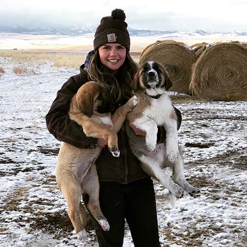Ashley Turner stood in the snow, wearing cold weather clothing including a beanie hat, whilst holding two dogs. There are hay bales in the background