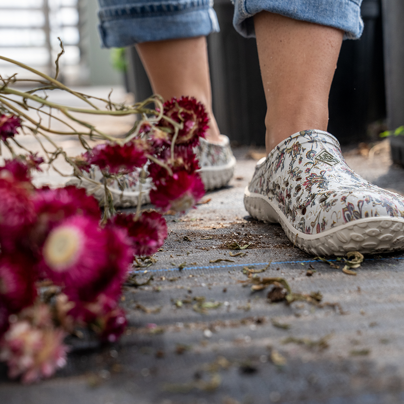 Close up image of a woman wearing a pair of floral print Muck Boot Muckster Lite Clogs, walking along a wooden floor with red flowers in the foreground