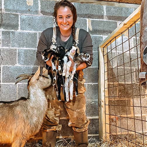 Elizabeth Taffet stood in a barn with breeze blocks behind her. She is holding four goat kids with the mother goat stood next to her. Elizabeth is wearing a long sleeve t-shirt, camouflage dungarees and a pair of Muck Boot Wellingtons  