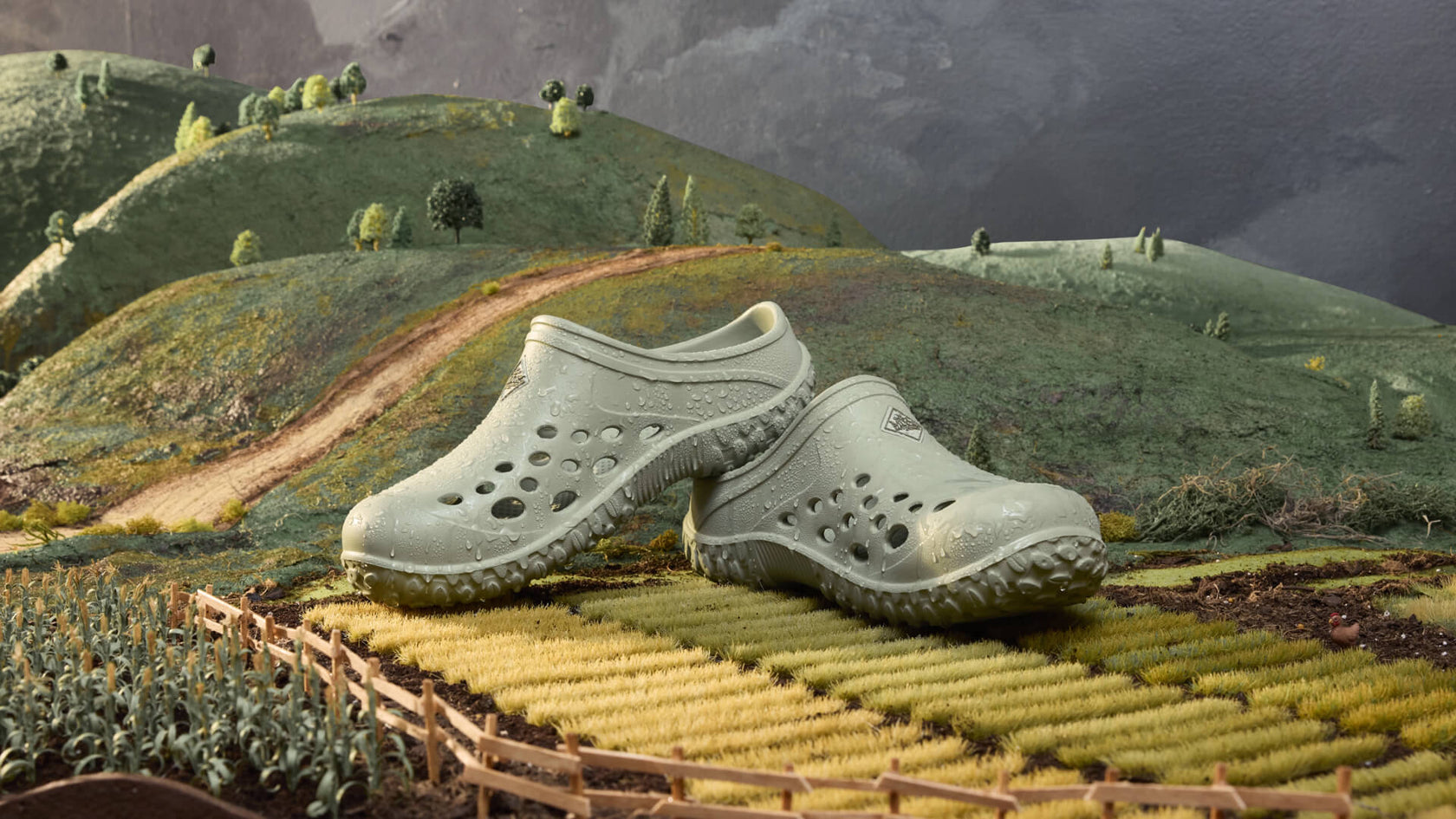Green Muck Boots Muckster clogs on a miniature landscape with hills and a path.