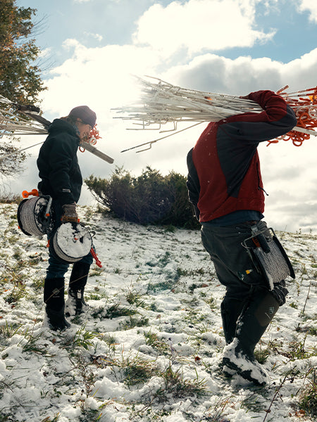 Two people walking in a snow covered field, carrying poles and wires