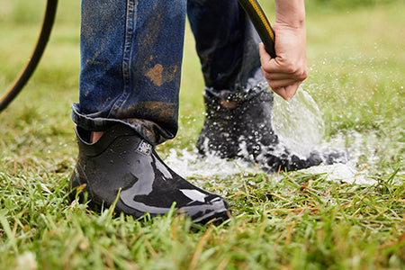 Close up of a person watering grass with a hosepipe, wearing a pair of black Muck Boot Wooster boots