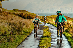 A woman and two children wearing Muck Boots and riding their bikes down a country lane 