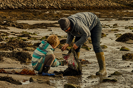 Adult and child collecting debris from a wet, sandy beach, both wearing cold weather clothing and a pair of Muck Boots