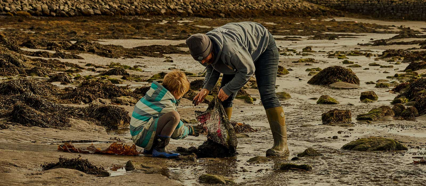 Adult and child collecting debris from a wet, sandy beach, both wearing cold weather clothing and a pair of Muck Boots