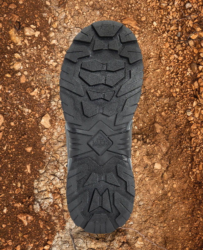 A Muck Boots mixed terrain outsole with a rocky and muddy texture behind