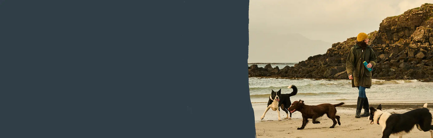 A woman wearing a yellow docker hat and Muck Boots wellingtons on a beach with three dogs.