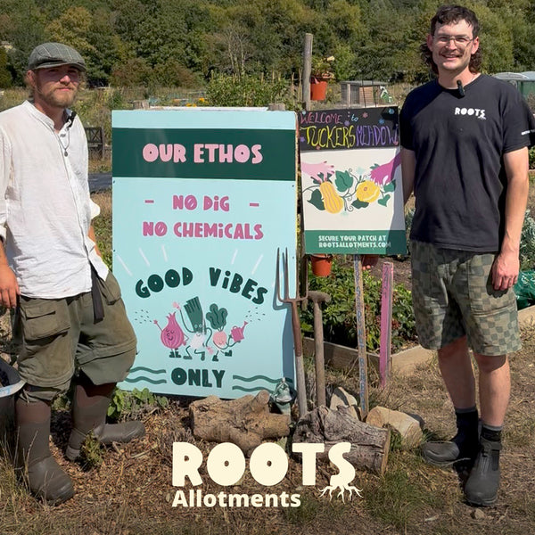 Two men standing in an allotment with a sign about their ethos and 'ROOTS Allotments' branding, wearing Muck Boots wellingtons.