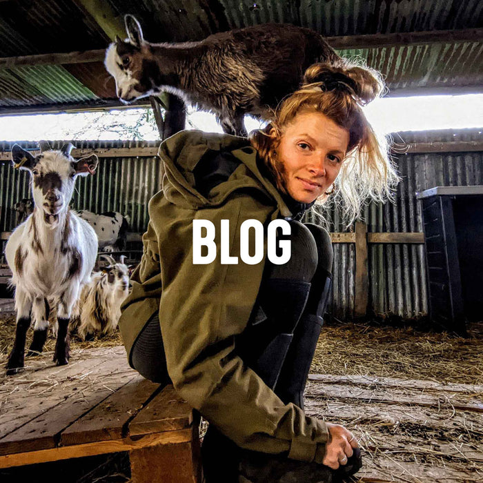 Zoë Colville sat on a wooden pallet in a barn, leaning over wearing a jacket and pair of Muck Boots with a goat stood on her back and others goats behind her. Text reads 'BLOG'