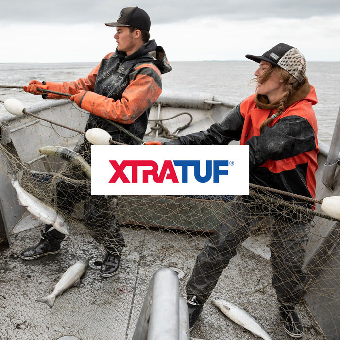A man and woman hauling fish onto a boat at sea. They're both wearing baseball caps, orange jackets and Xtratuf Ankle Deck Boots