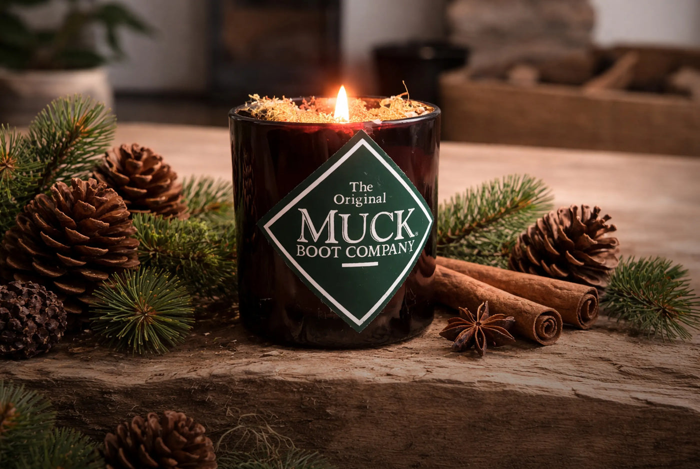 A lighted candle branded with the Muck Boots logo surrounded by pine cones and needles on a wooden bench.