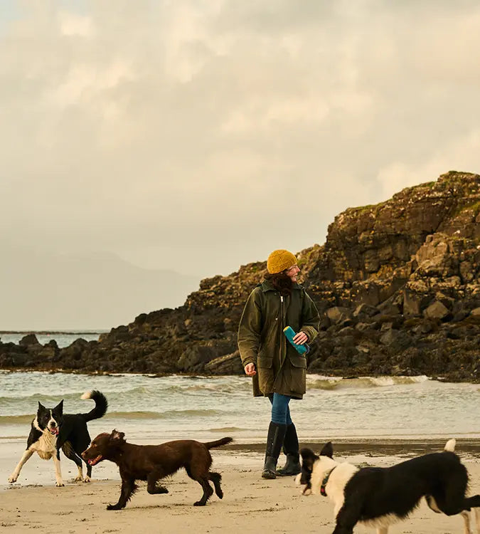 A woman wearing a yellow docker hat and Muck Boots wellingtons on a beach with three dogs.
