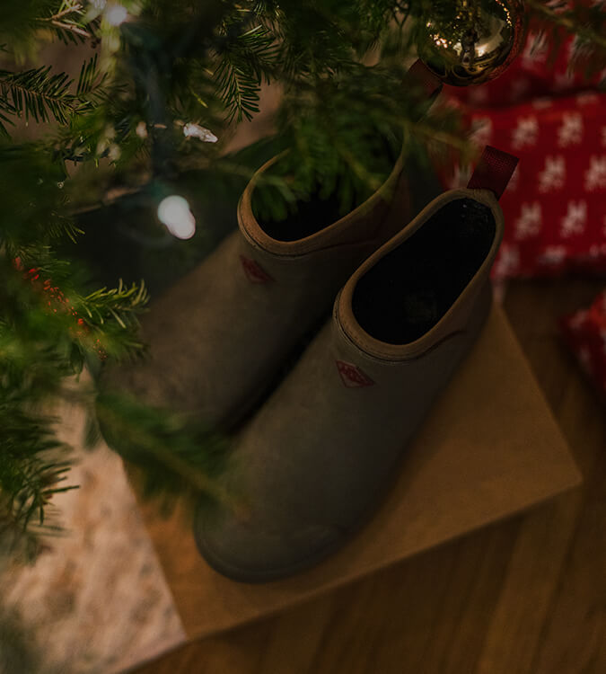 A pair of Muck Boots amongst a Christmas tree with lights and wrapped presents.