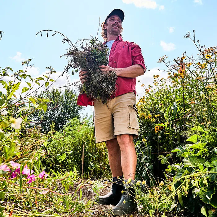 A man wearing a pair of Muck Boots Rainscape boots, holding a bunch of dead plants and surrounded by wildflowers.