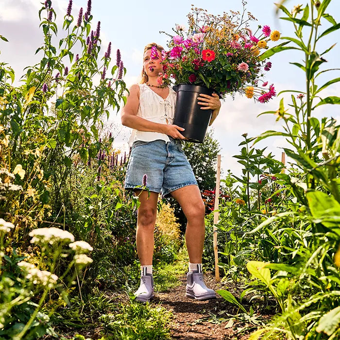 A woman wearing Muck Boots Rainscape boots holding a large plastic pot filled with flowers.