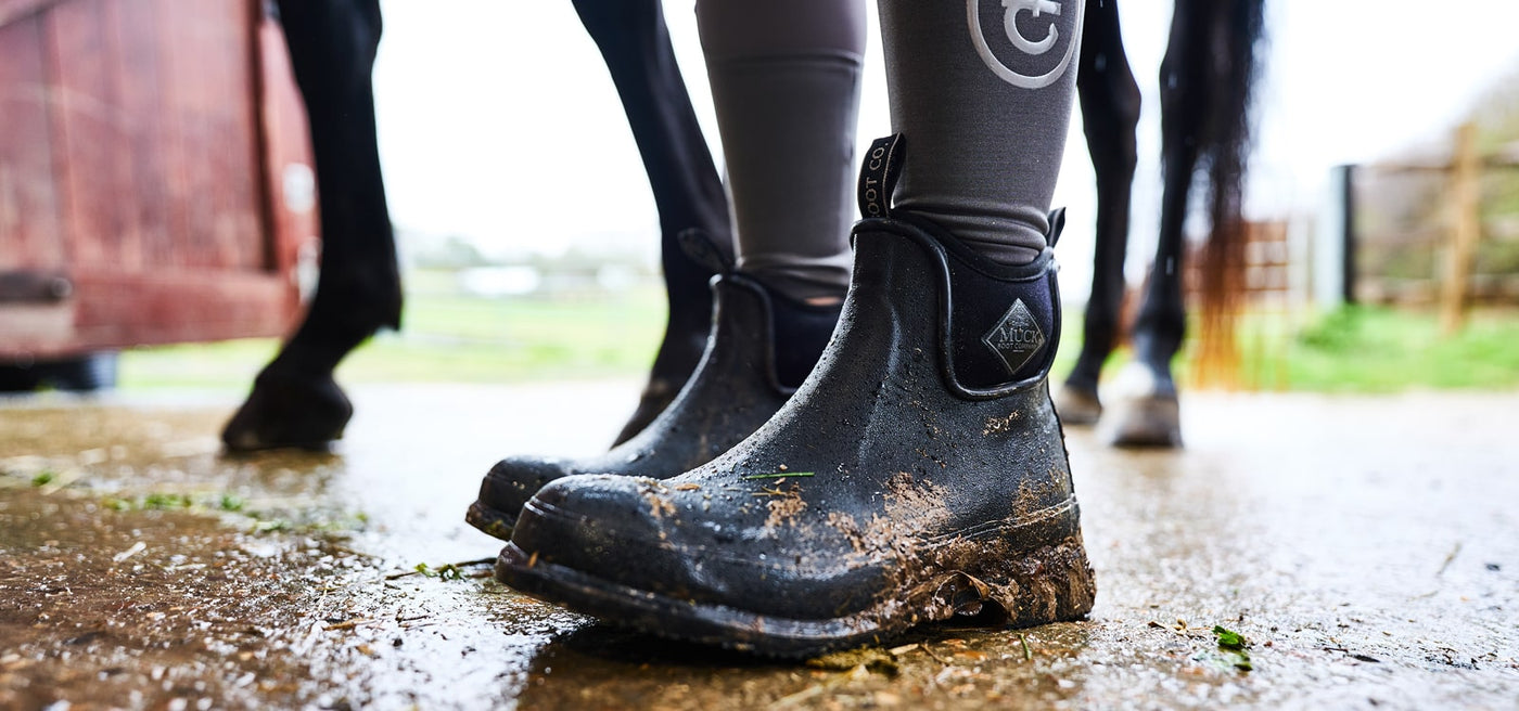 Close up image of a person wearing a pair of muddy Muck Boots Unisex Wear Ankle Boots in a horse stable