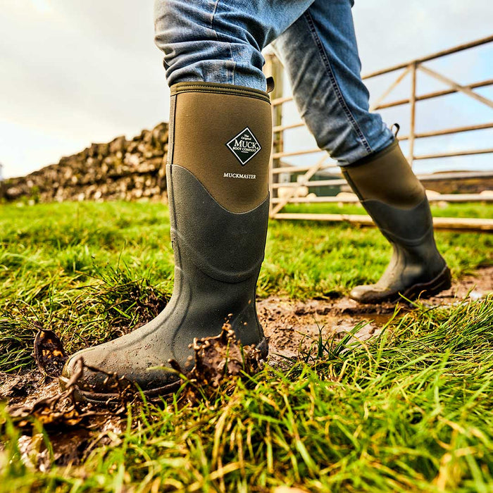 Close up of a person walking away from a dry stone wall and metal gate, through a muddy field, wearing a pair of Muck Boots Muckmaster Wellingtons 