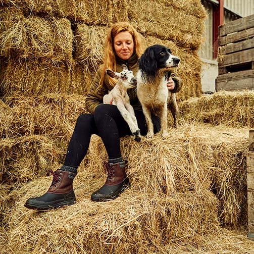 Zoe Colville sat on a haybale with a lamb and dog, wearing a pair of Muck Boots