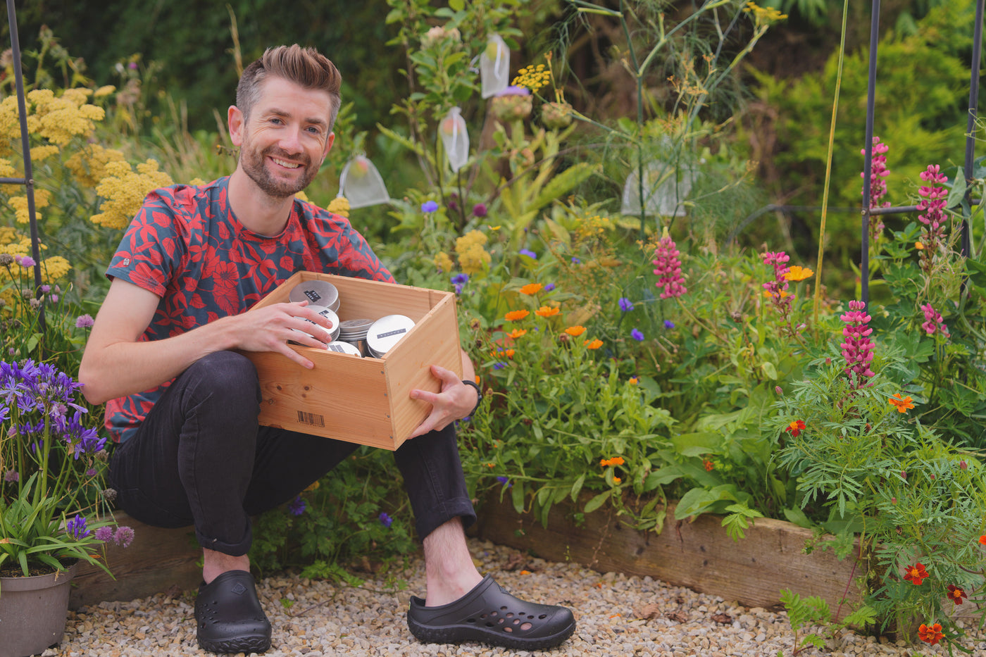 Niall sat by a flower bed holding a wooden box with metal tins containing seeds inside. Niall is wearing a pair of Muck Boots Muckster Lite Clogs in black