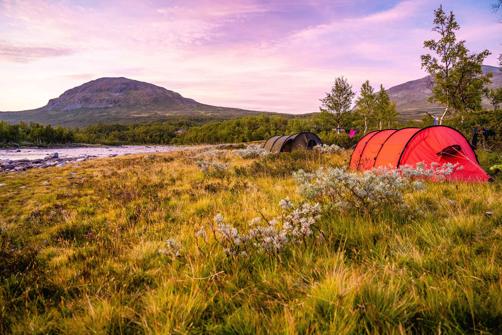 Two camping tents amongst a picturesque field with a river running through and mountains in the background