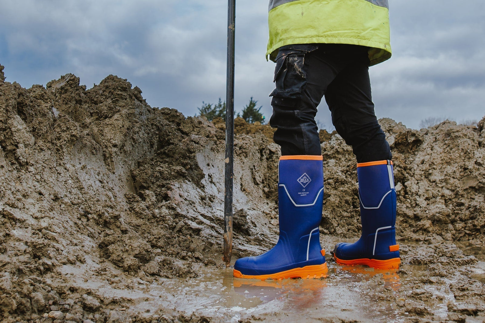 A constructor working surveying a muddy building site, wearing a yellow hi-vis jacket and a pair of blue Muck Boots Grit Safety Boots