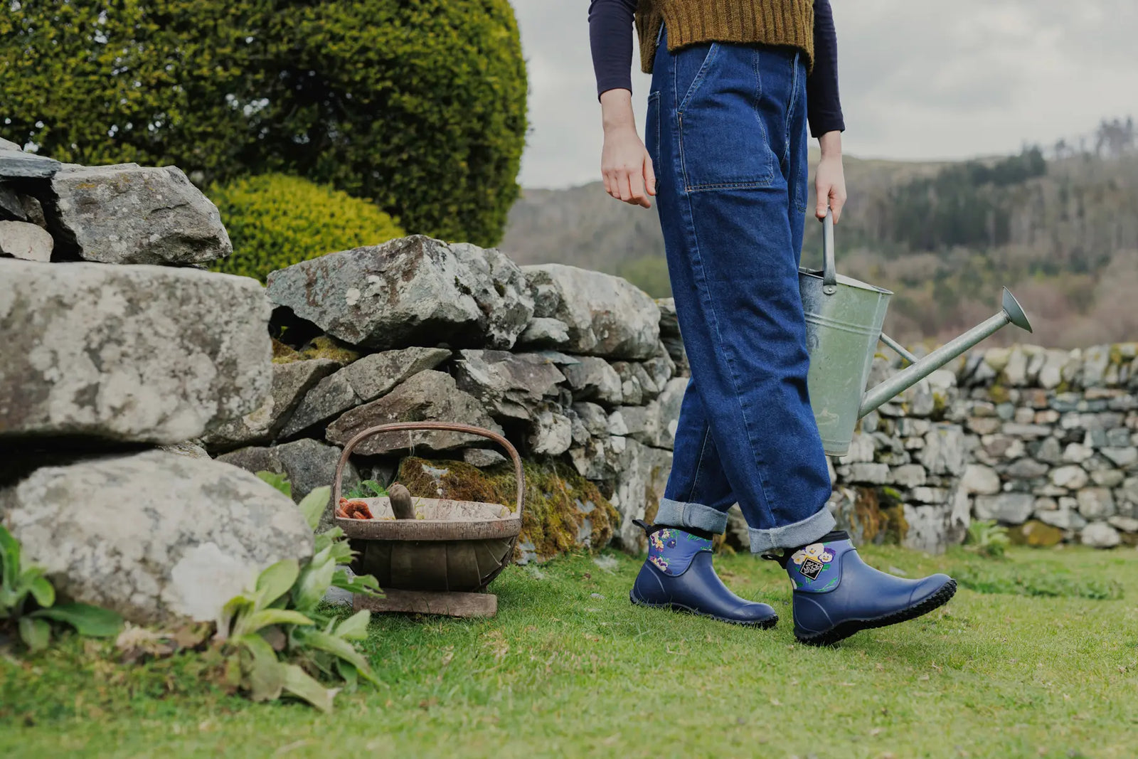Woman stood in front of a dry stone wall, holding a watering can and wearing a pair of Muck Boots RHS Muckster II Ankle Boots