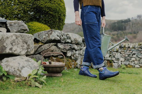 Woman stood in front of a dry stone wall, holding a watering can and wearing a pair of Muck Boots RHS Muckster II Ankle Boots