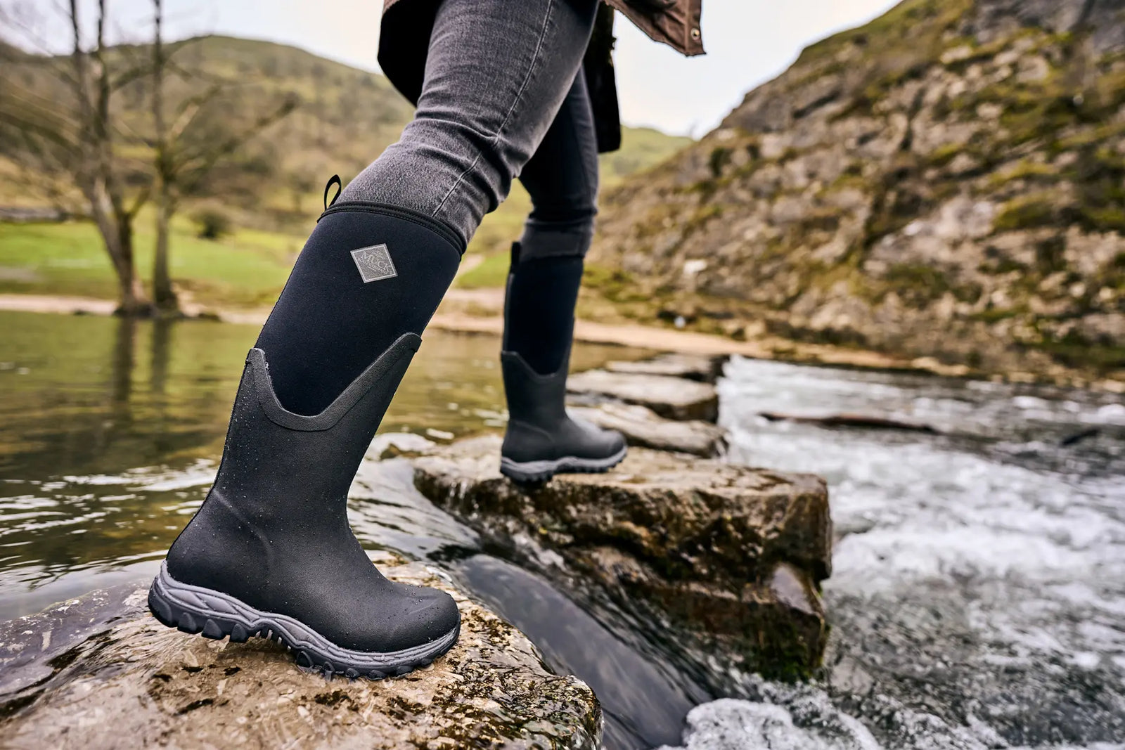 Person stepping between wet rocks in a river, wearing a pair of Muck Boots Arctic Sport II wellingtons