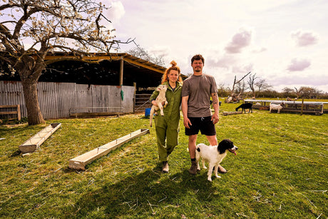 Zoe Colville and her husband outside on a farm with Zoe holding a lamb and a dog standing in front of her husband.