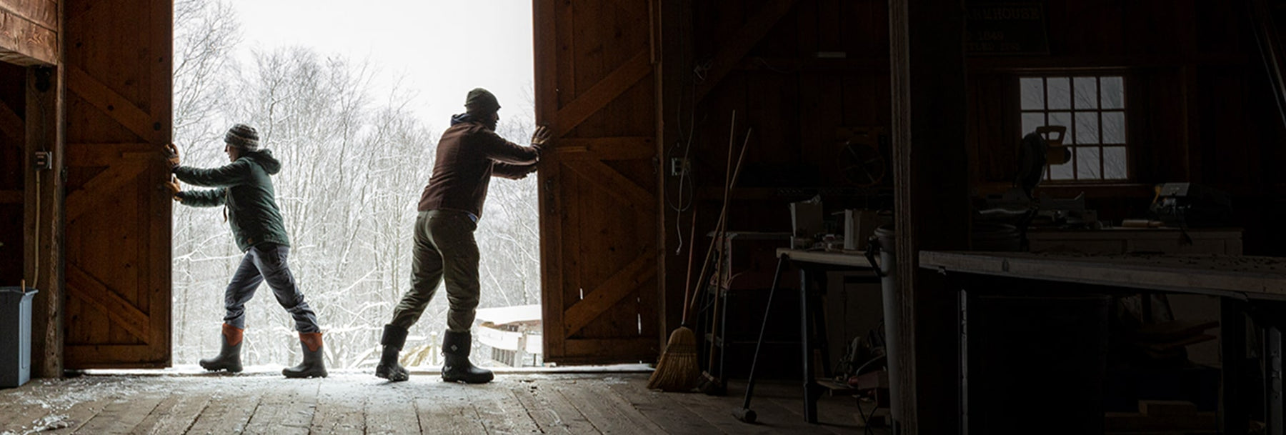 Two people wearing Muck Boot wellingtons, pushing barn doors open with a snowy scene outside