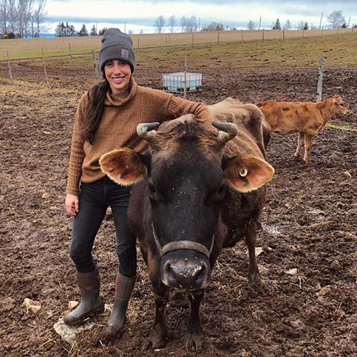 Ashley Turner stood in a muddy field with a cow and calf. She is wearing a beanie hat, brown jumper, jeans and a pair of Muck Boots