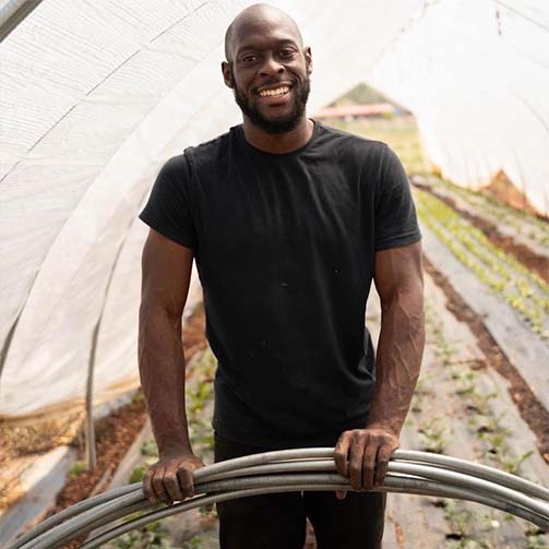 Kamal Bell stood in a polytunnel, wearing a black t-shirt with rows of crops behind him