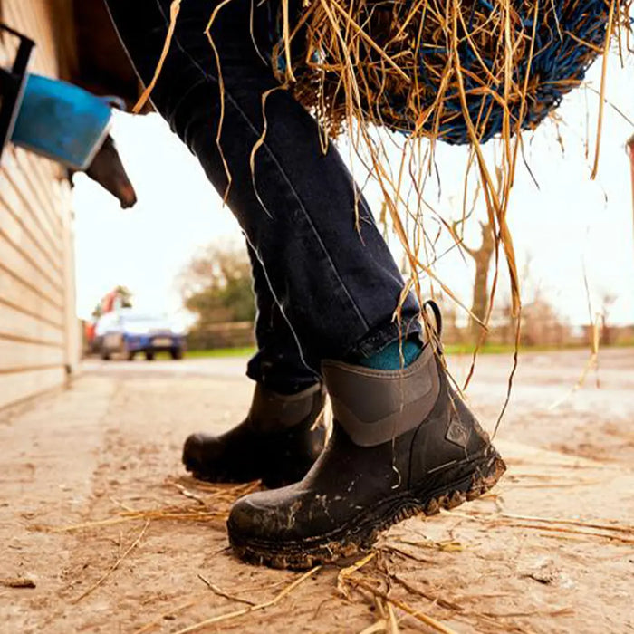 Close-up image of a person in a yard wearing a pair of Muck Boots Women's Arctic Sport II Ankle Boots, carrying hay.