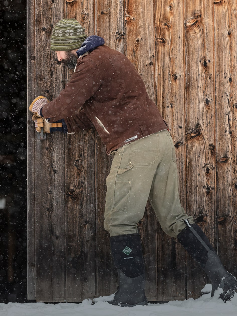 Close-up image of a man wearing a pair of Muck Boots Arctic Sport Wellingtons in the snow outside of a n open wooden barn door