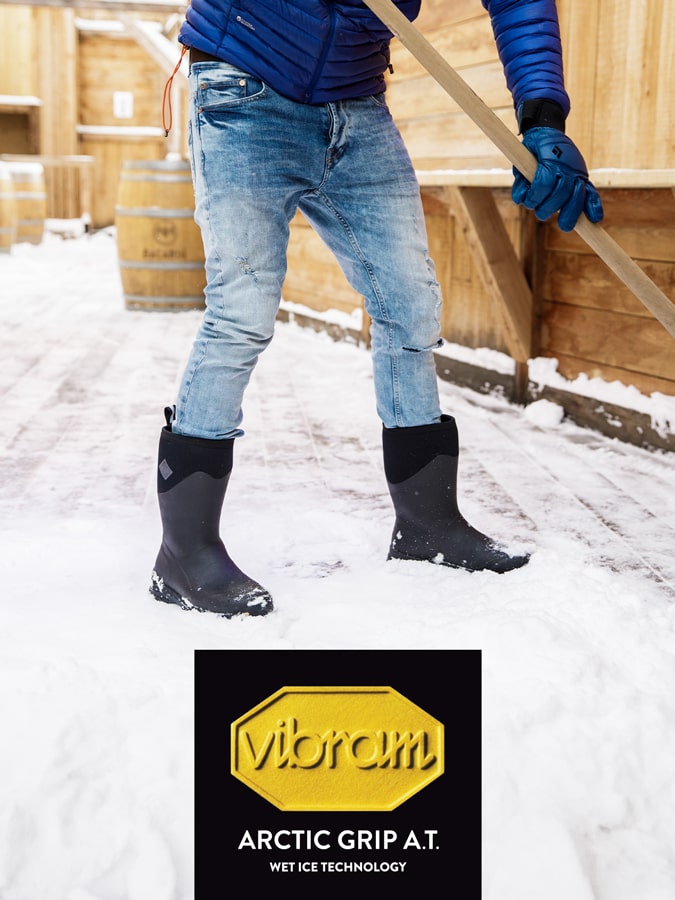 Close-up image of a person stood shovelling snow, wearing a pair of Muck Boots. Vibram logo at the bottom of image.