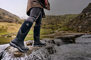 Close up of a woman wearing a pair of Muck Boot Arctic Sport II Wellingtons, stood on a stepping stone in a river with flowing water 