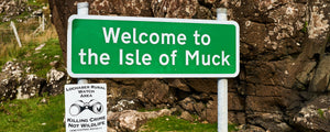 A green sign that has 'Welcome to the Isle of Muck' written on it in front of rocks and boulders