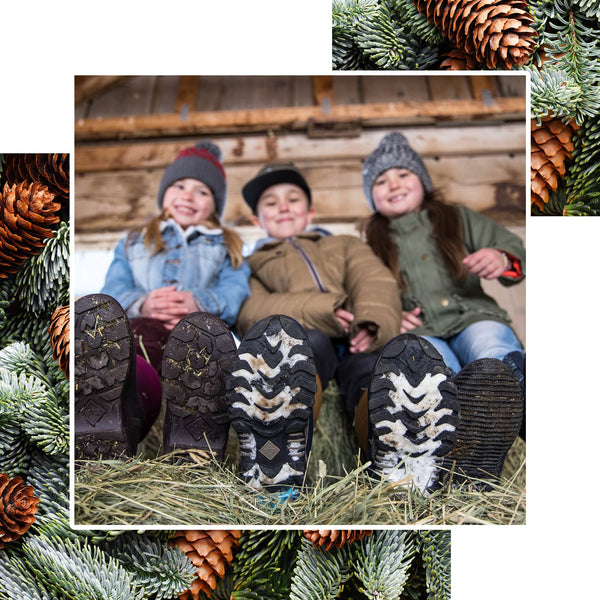 Three children sat in a barn wearing cold weather clothing and Muck Boots wellingtons with a pine tree texture border around the image