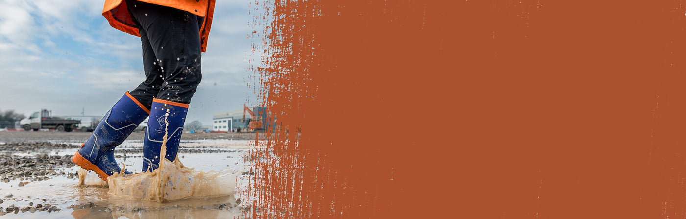 Person walking through a wet and muddy building site wearing a pair of blue Muck Boots Grit S5 Wellingtons