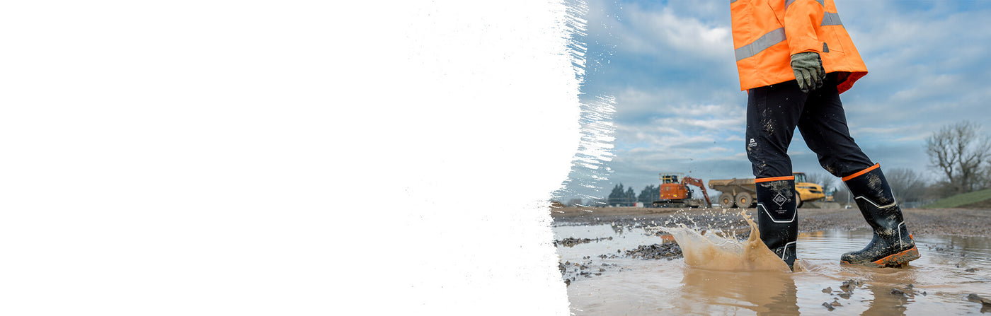 A man walking through muddy puddles on a building site, wearing a pair of Muck Boots Grit Safety Wellingtons