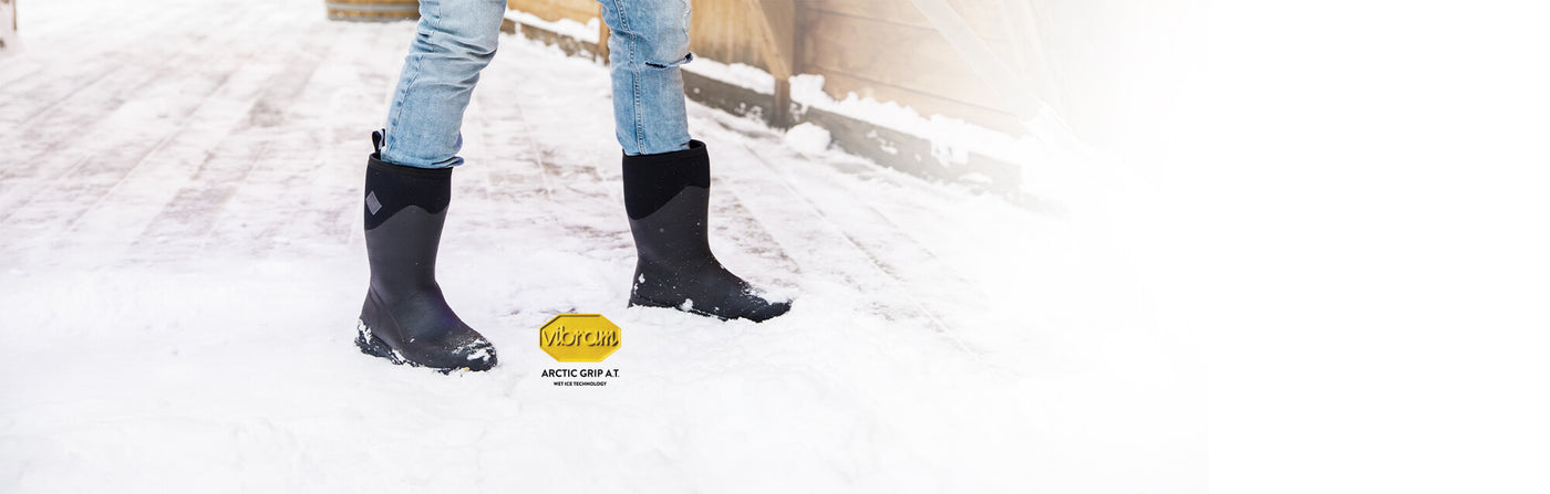 Person shovelling snow, wearing a pair of Muck Boots Arctic Ice Vibram wellingtons with a Vibram logo to the left of the image