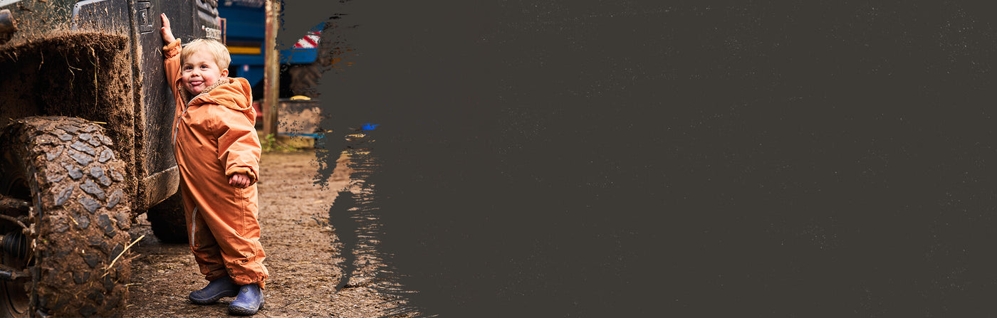 Child wearing a pair of blue Muck Boots  Kids Muckster Boots in an orange jumpsuit standing next to a large tire on a dark background