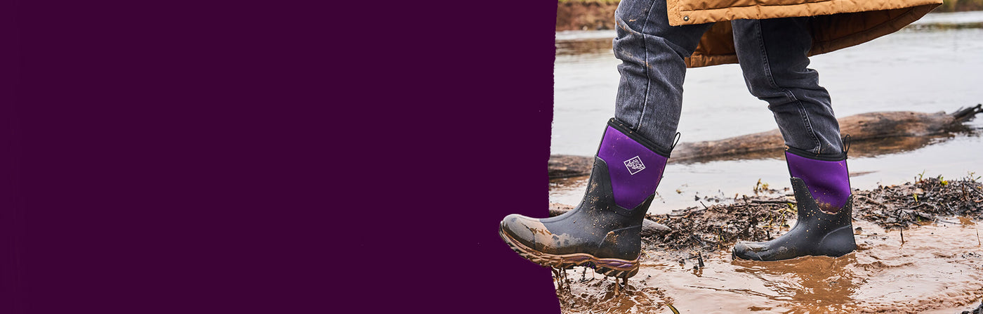 Person wearing a pair of Muck Boots Women's Arctic Sport II boots in black acai, walking through a muddy puddle.