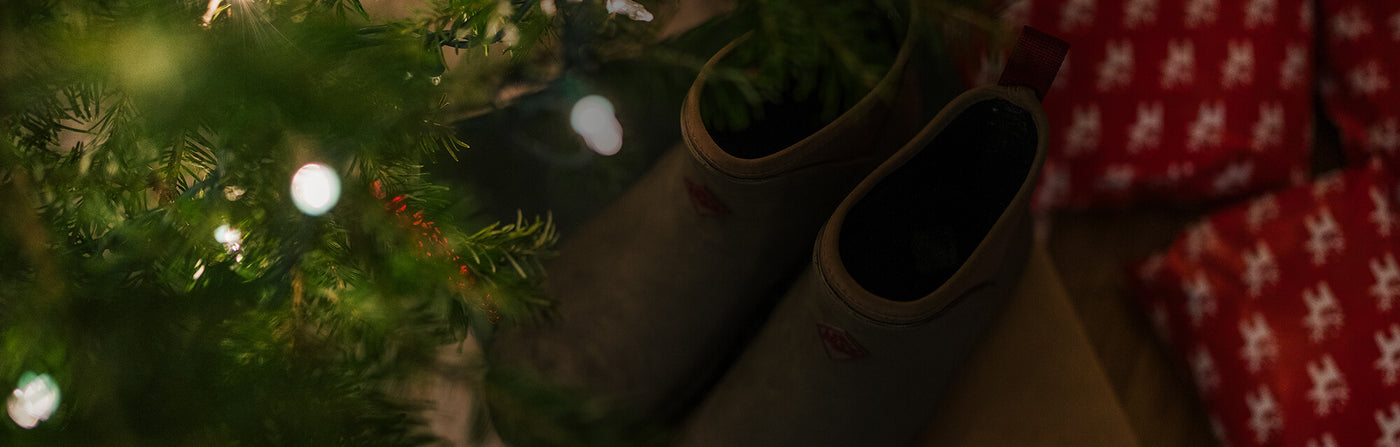 A pair of Muck Boots amongst a Christmas tree with lights and wrapped presents.