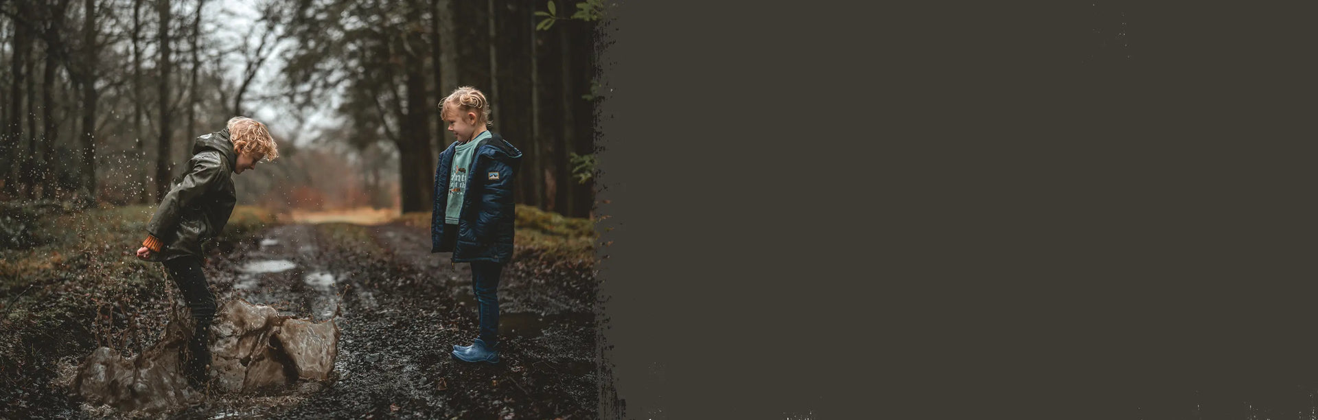Two children wearing Muck Boots wellingtons on a muddy lane, with one jumping in a puddle.