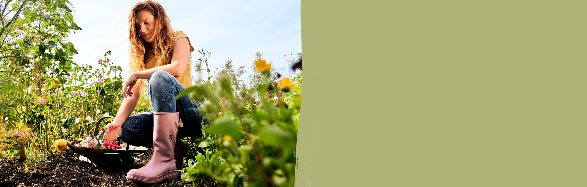 Woman kneeling down amongst wildflowers with a pair of secateurs in her hand, wearing a pair of Woodrose twilight mauve Muck Boots Tall Rainscape Wellingtons. 