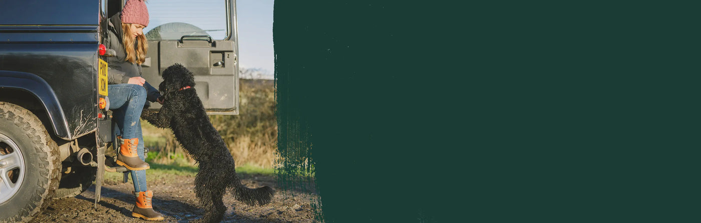A woman wearing a pair of Tan Muck Boots Muck Originals Duck Lace-Up Leather Short Boots, sat in the back of a Land Rover feeding a black dog.
