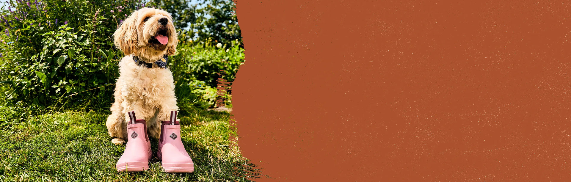 A dog wearing a pair of Muck Boots Women's Rainscape Chelsea Boots in Woodrose twilight mauve outside surrounded by lush greenery and sunshine.